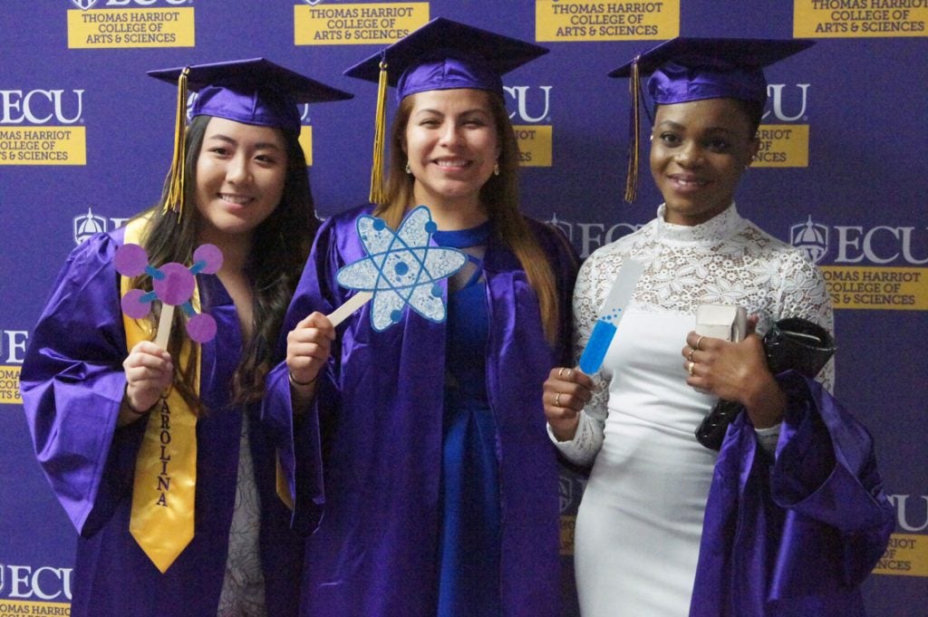 Three undergraduate women in graduation regalia holding paper cutouts of a molecule, atom, and test tube.