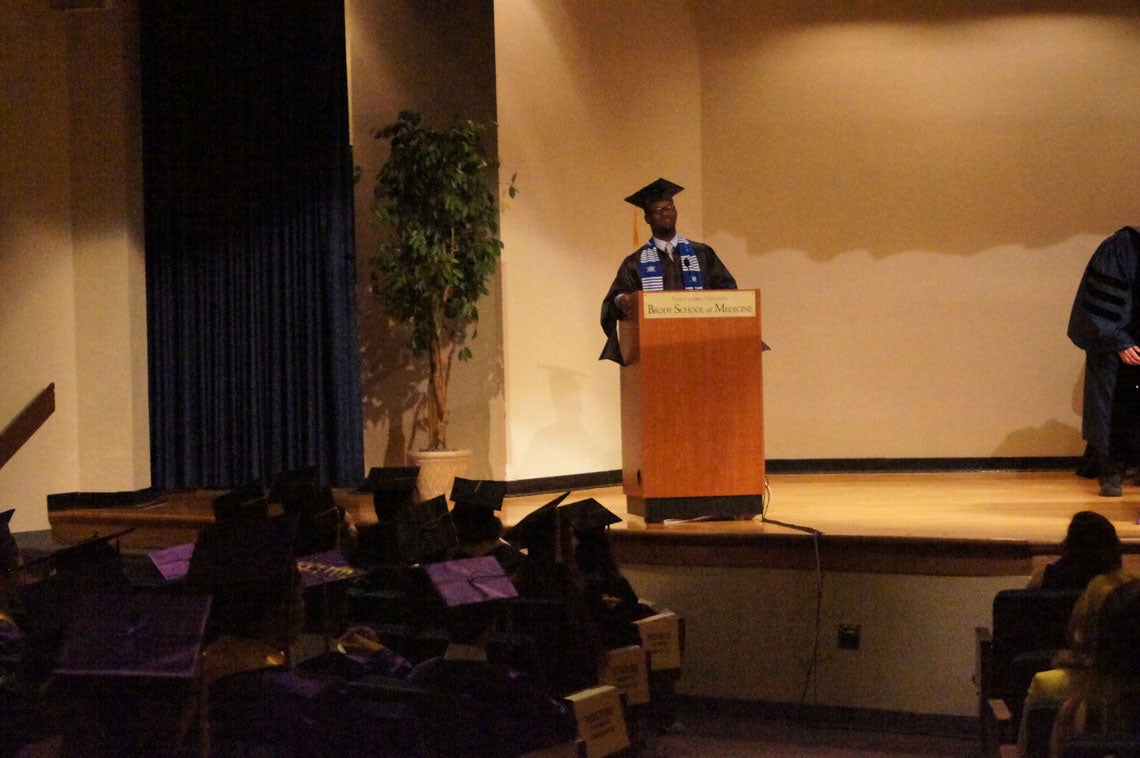African American man standing behind a podium speaking to the graduating class.
