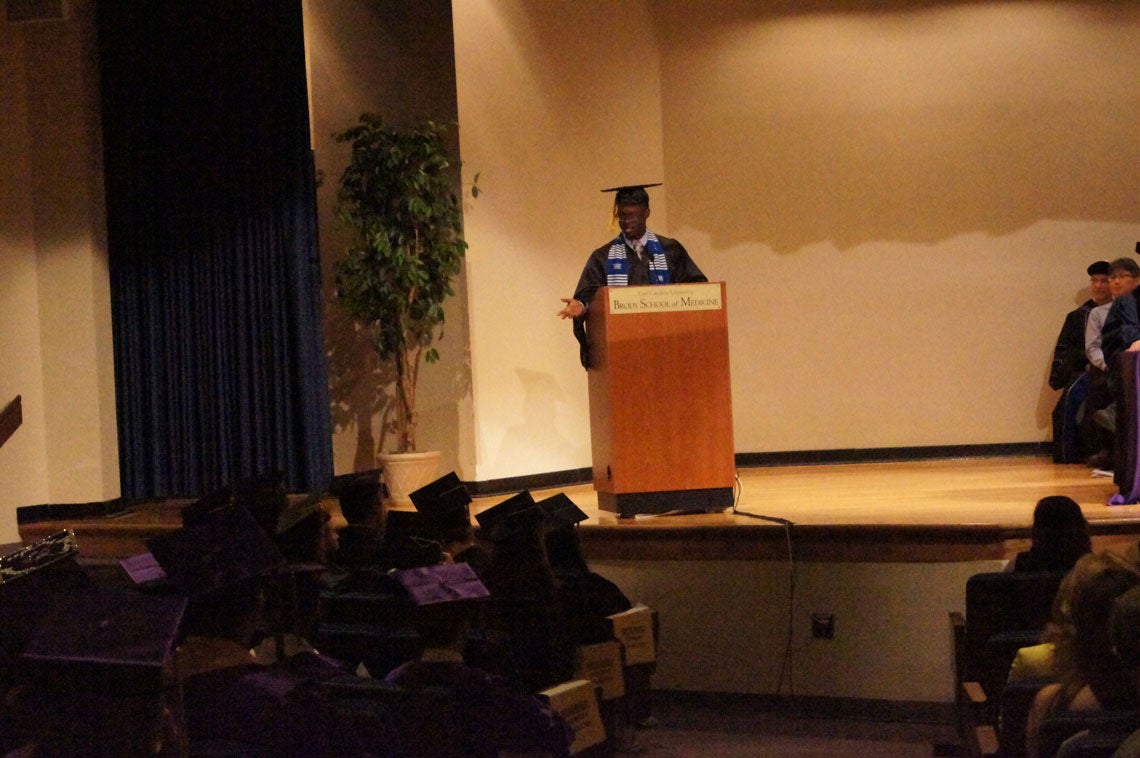 African American man standing behind a podium speaking to the graduating class.