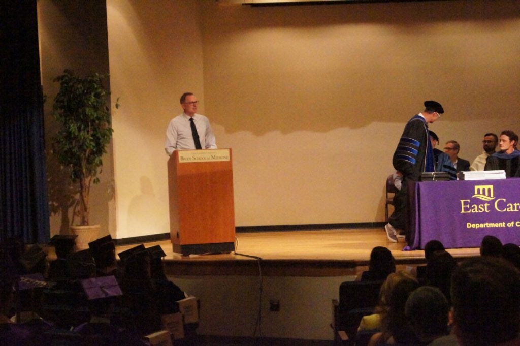 White man standing behind a podium on stage at a graduation ceremony.