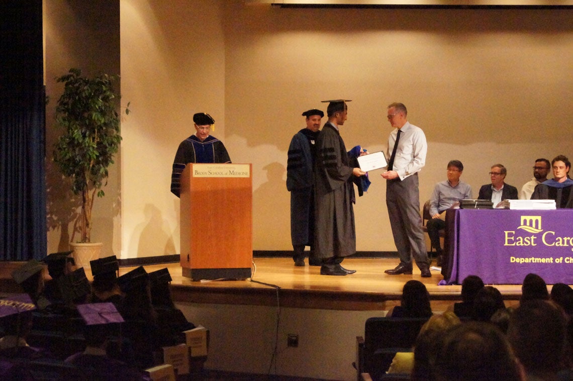 A graduate student shakes hands with faculty members on stage at graduation.