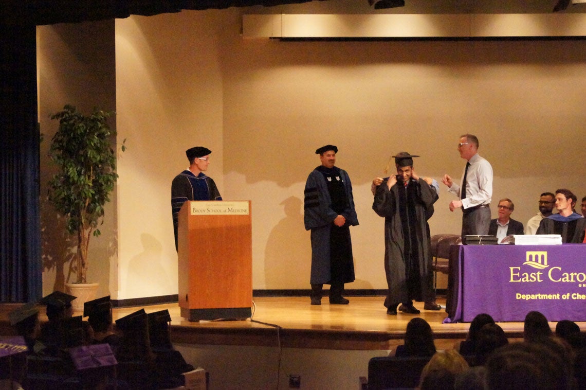 A graduate student receives his hood on stage at a graduation ceremony.