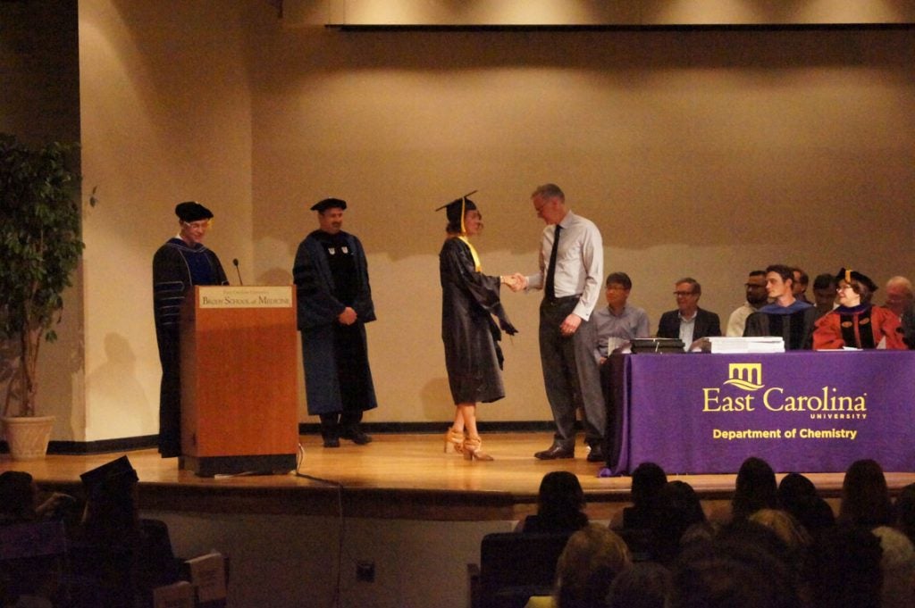 A graduate student shakes hands with faculty on stage at a graduation ceremony.
