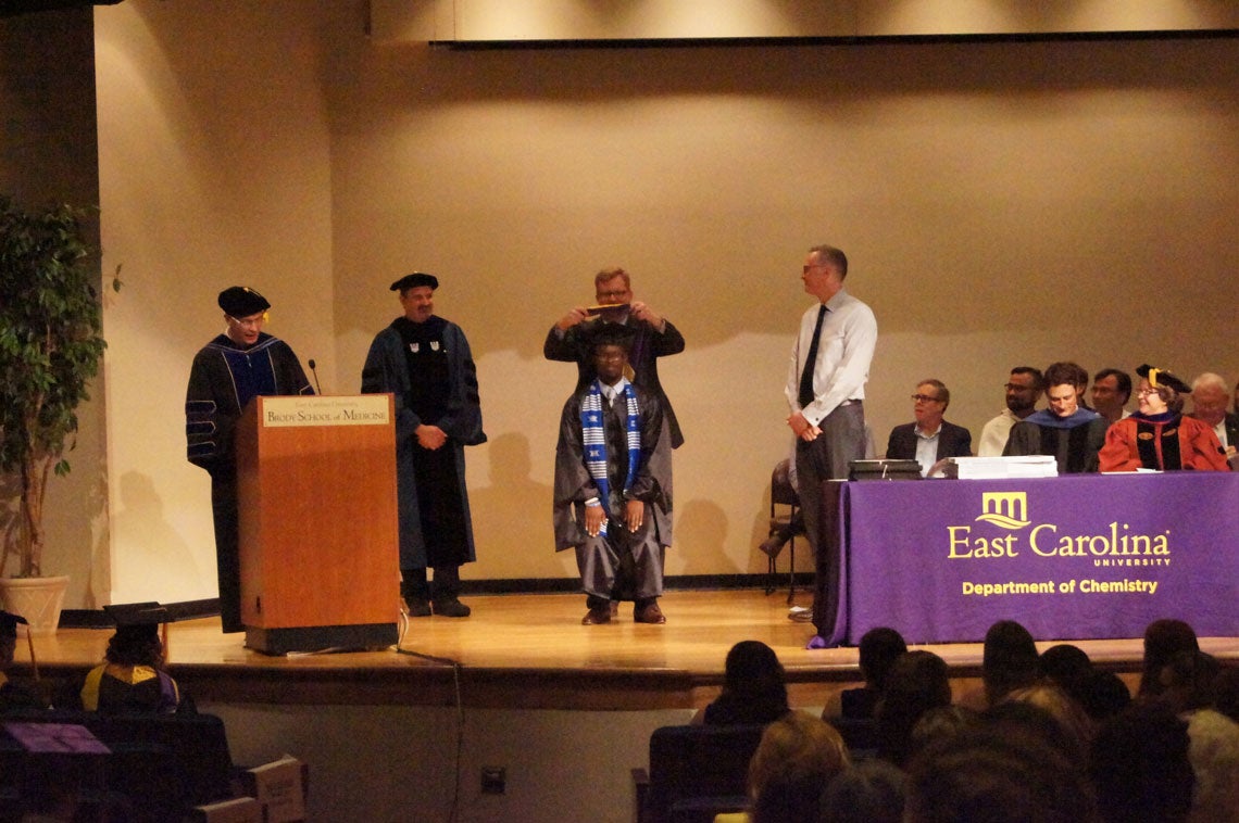 A graduate student receives his hood on stage at a graduation ceremony.