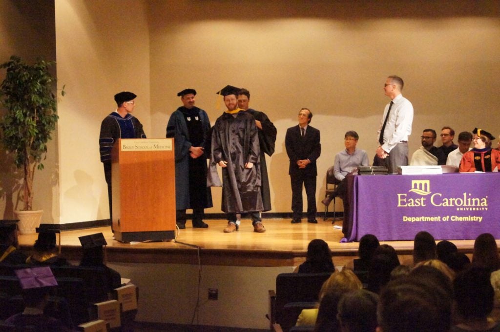 A graduate student receives his hood on stage at a graduation ceremony.