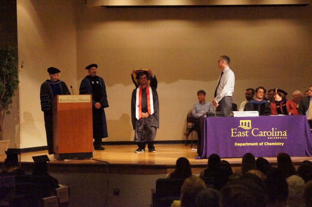 A graduate student receives his hood on stage at a graduation ceremony.