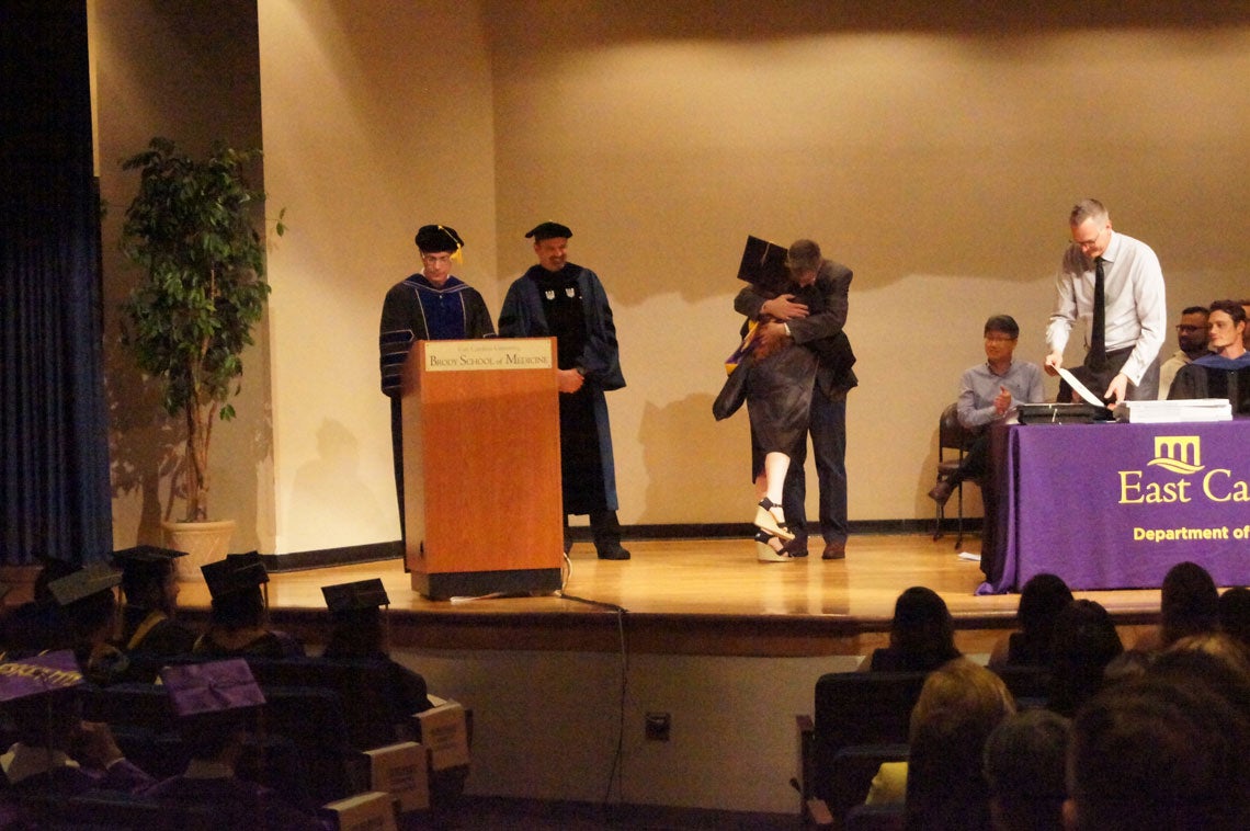A graduate student hugs a man on stage at a graduation ceremony.