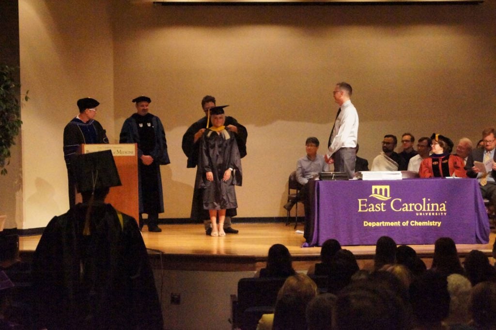 A graduate student receives her hood on stage at a graduation ceremony.