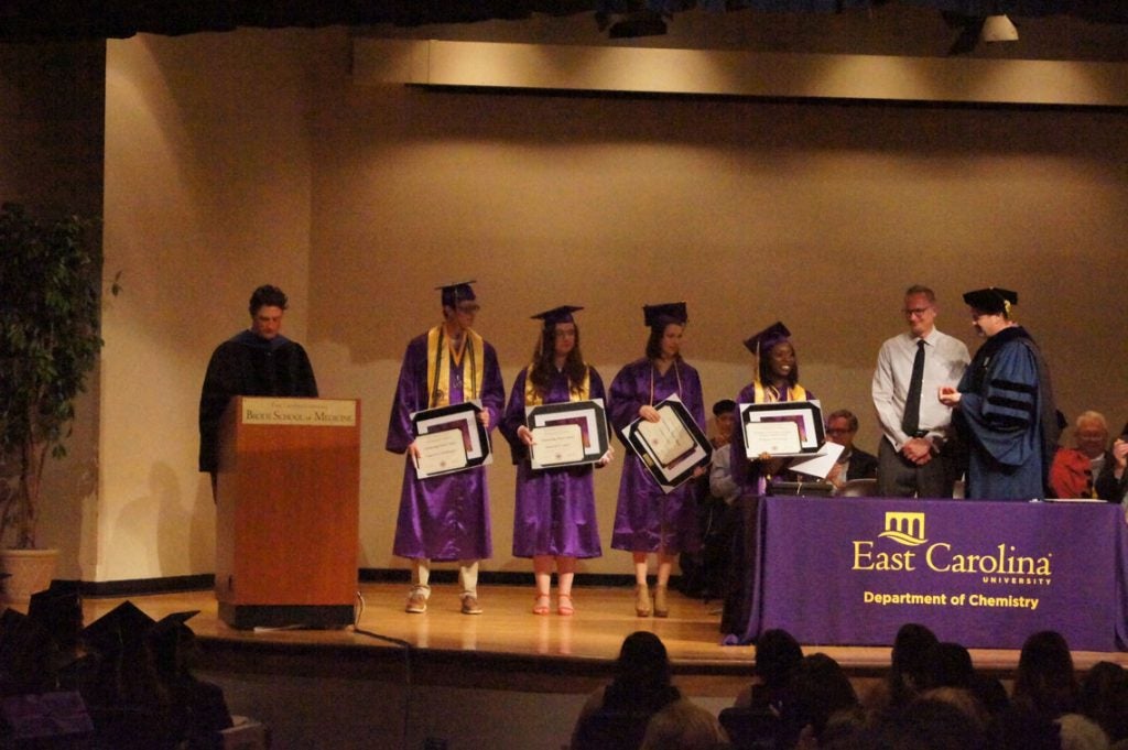 Four undergraduate students stand on stage with their degrees and a framed item.