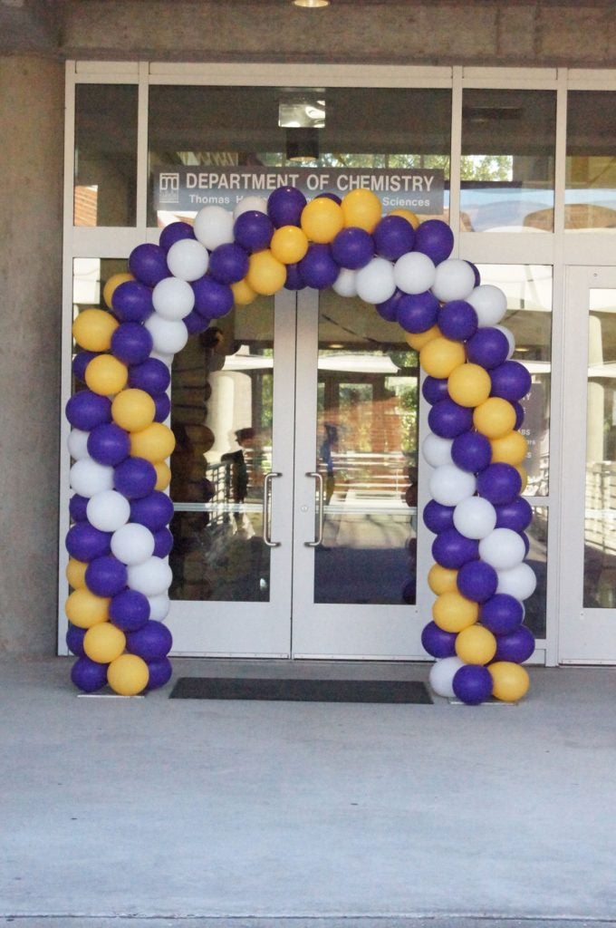 Purple, gold and white baloon arch over doorway with a Department of Chemistry sign over it.