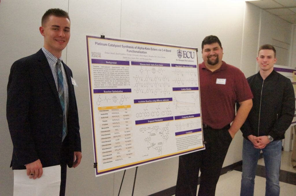 Three male students standing next to a poster about Platinu Catalyzed Synthesis of Alpha-Keto Esters via C-H bond Functionalization.