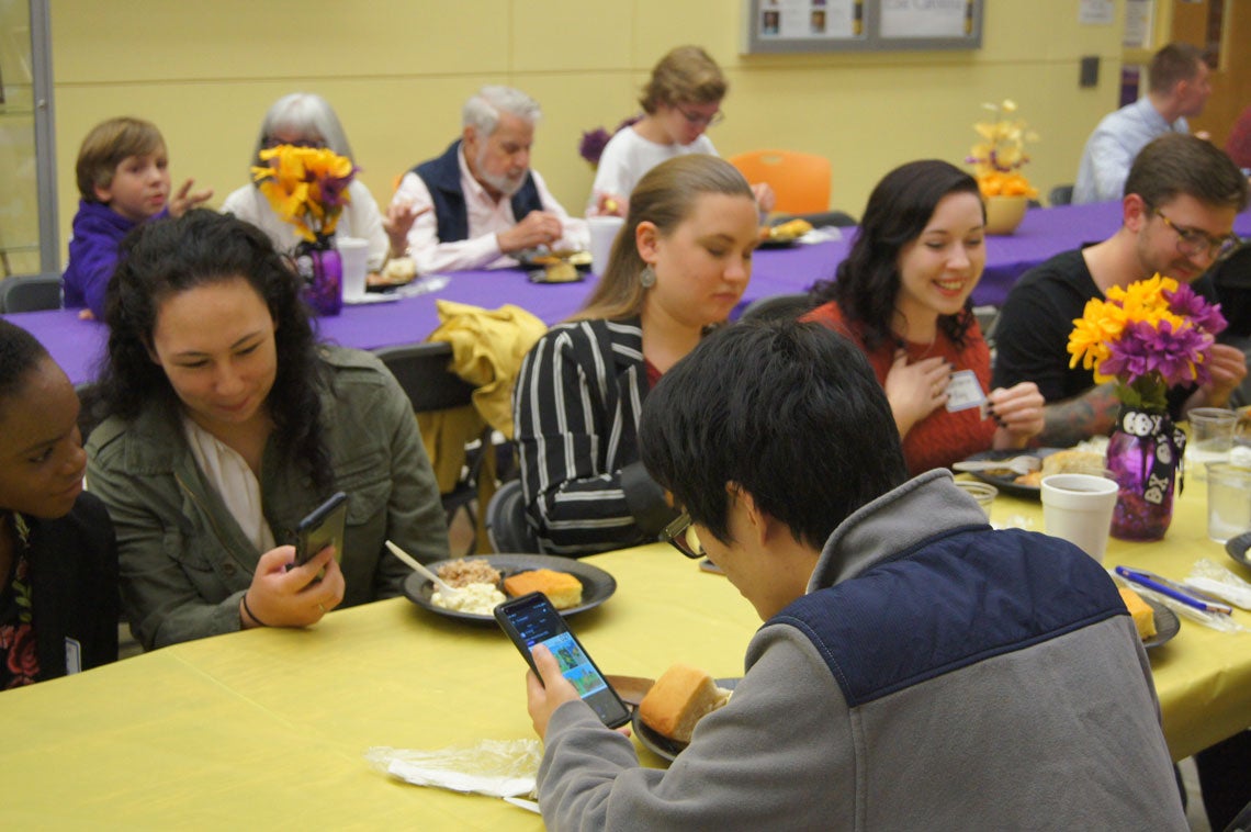 Group of people eating at long tables with purple and gold tablecloths and centerpieces.