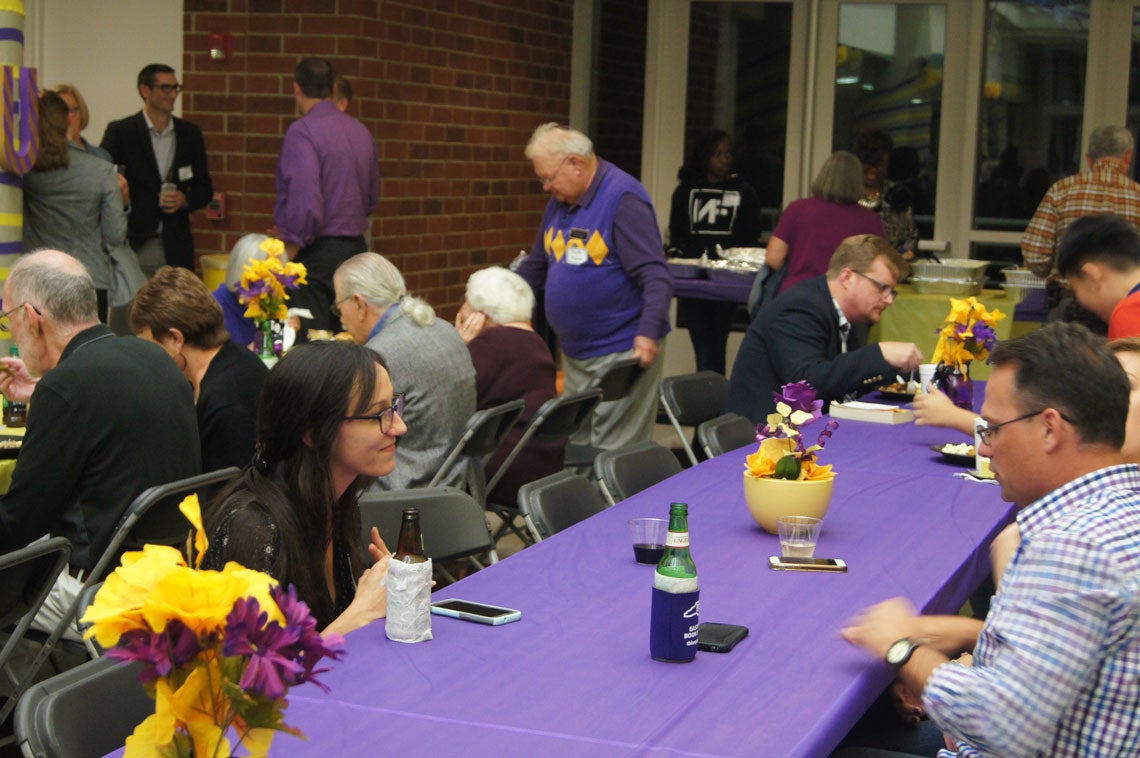 Group of people eating and mingling around long tables with purple and gold tablecloths and centerpieces.