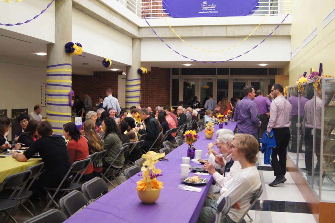 Group of people eating and mingling around long tables with purple and gold tablecloths and centerpieces.