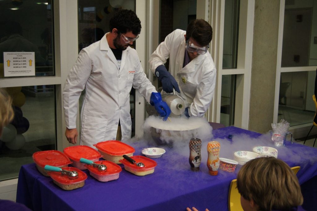 Two men in lab coats and goggles making ice cream with liquid nitrogen.