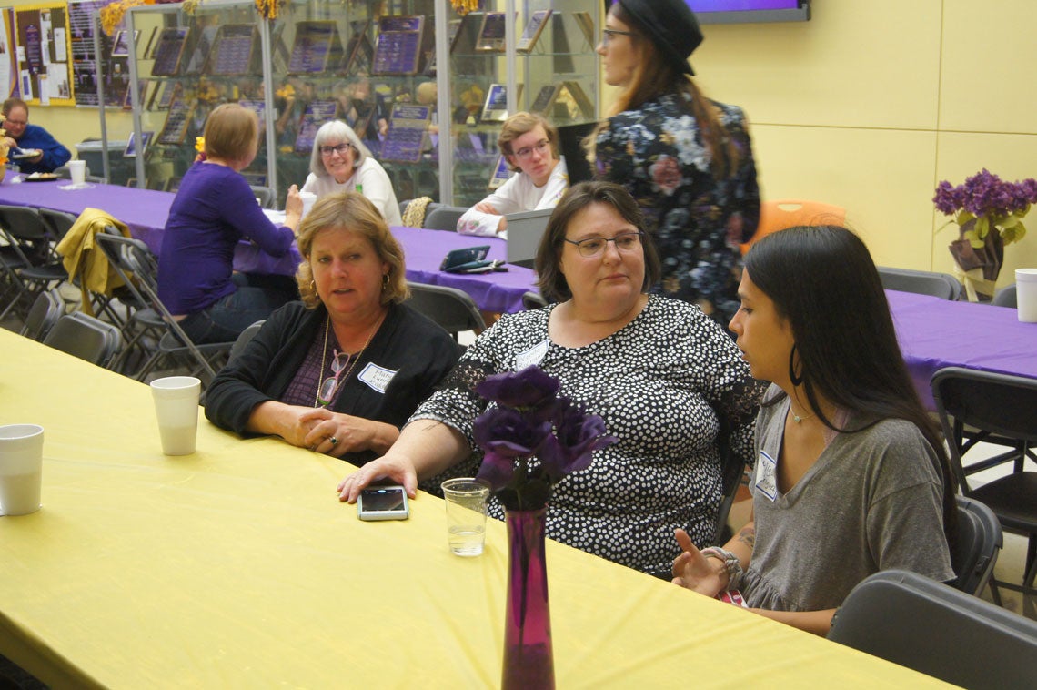 Group of people mingling around long tables with purple and gold tablecloths and centerpieces.