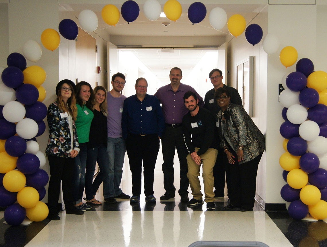Group of people standing underneath a purple, gold and white balloon arch.